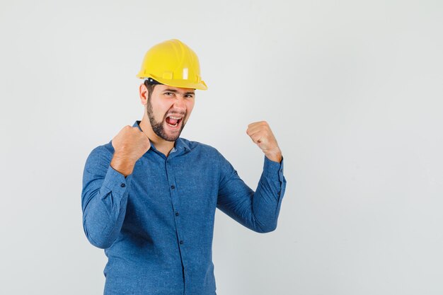 Joven trabajador en camisa, casco mostrando gesto de ganador y mirando feliz