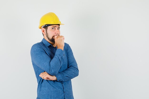 Joven trabajador en camisa, casco de pie en pose de pensamiento y mirando asustado