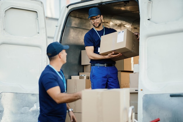 Joven trabajador cargando cajas de cartón en una furgoneta de reparto y comunicándose con su colega