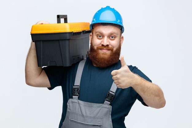 Joven trabajador de la construcción complacido con casco de seguridad y uniforme sosteniendo una caja de herramientas en el hombro mirando a la cámara mostrando el pulgar hacia arriba aislado en el fondo blanco