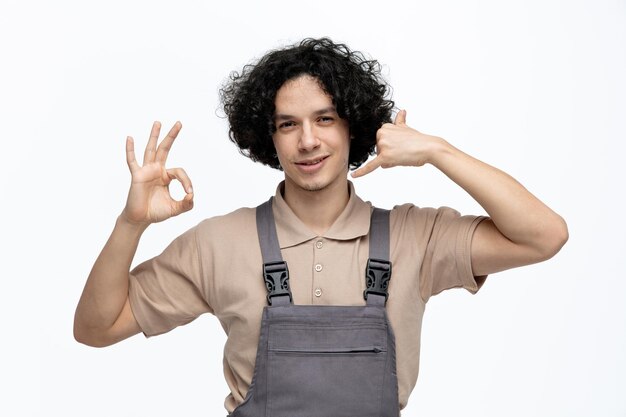 Joven trabajador de la construcción complacido con uniforme mirando a la cámara mostrando el signo de estar bien y llámame gesto aislado en el fondo blanco