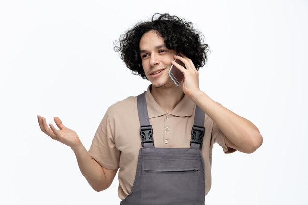 Un joven trabajador de la construcción complacido con uniforme que muestra la mano vacía mirando a un lado mientras habla por teléfono aislado de fondo blanco