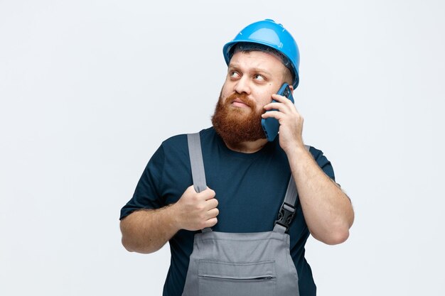 Joven trabajador de la construcción confiado usando casco de seguridad y uniforme agarrando su uniforme mirando hacia arriba mientras habla por teléfono aislado en fondo blanco