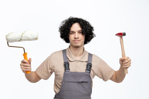 Un joven trabajador de la construcción descontento con uniforme que sostiene un rodillo de pintura y un martillo mirando a la cámara aislada de fondo blanco