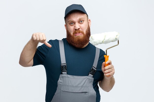 Un joven trabajador de la construcción disgustado con gorra y uniforme mirando a la cámara sosteniendo un rodillo de pintura mostrando el pulgar hacia abajo aislado en el fondo blanco