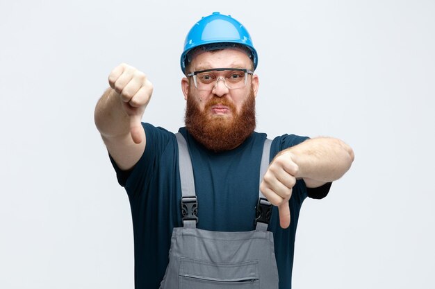Joven trabajador de la construcción disgustado con uniforme de casco de seguridad y gafas de seguridad mirando a la cámara mostrando los pulgares hacia abajo aislado en fondo blanco