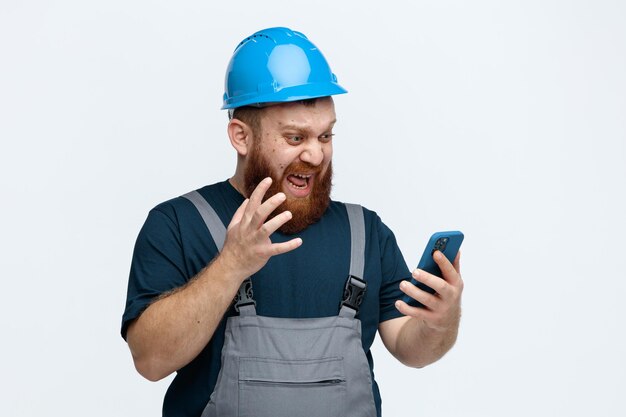 Joven trabajador de la construcción enojado con casco de seguridad y uniforme sosteniendo y mirando el teléfono móvil manteniendo la mano en el aire aislada en el fondo blanco