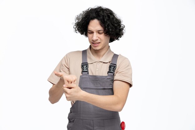 Joven trabajador de la construcción irritado con uniforme estirando los dedos mirándolos con instrumentos de construcción en el bolsillo aislado de fondo blanco