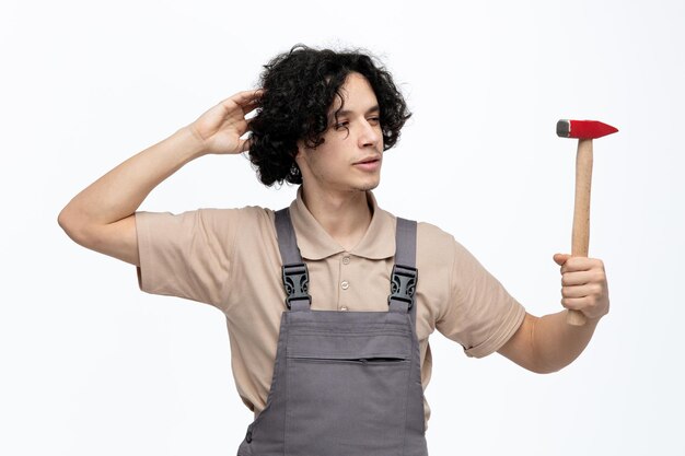 Un joven trabajador de la construcción pensativo con uniforme sosteniendo y mirando un martillo rascándose la cabeza aislado de fondo blanco