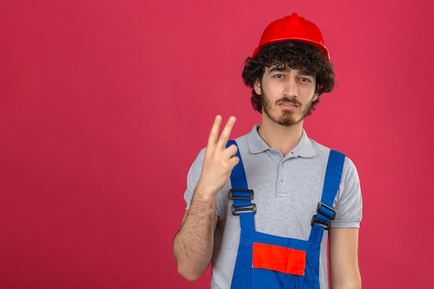 Joven trabajador constructor guapo empanado disgustado vistiendo uniforme de construcción y casco de seguridad mostrando y apuntando hacia arriba con los dedos número dos sobre fondo rosa aislado