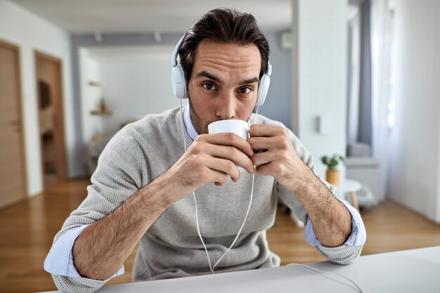 Joven trabajador independiente bebiendo café mientras escucha música con auriculares en casa