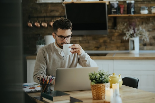 Foto gratuita joven trabajador independiente bebiendo café mientras trabaja en una computadora portátil en casa