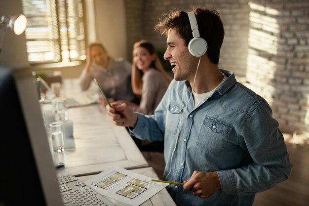 Joven trabajador independiente feliz escuchando música con auriculares y divirtiéndose mientras trabaja en la oficina Sus colegas están en segundo plano