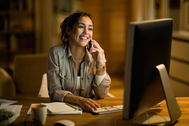 Joven trabajador independiente feliz que se comunica por teléfono móvil mientras navega por la red en una PC de escritorio por la noche en casa