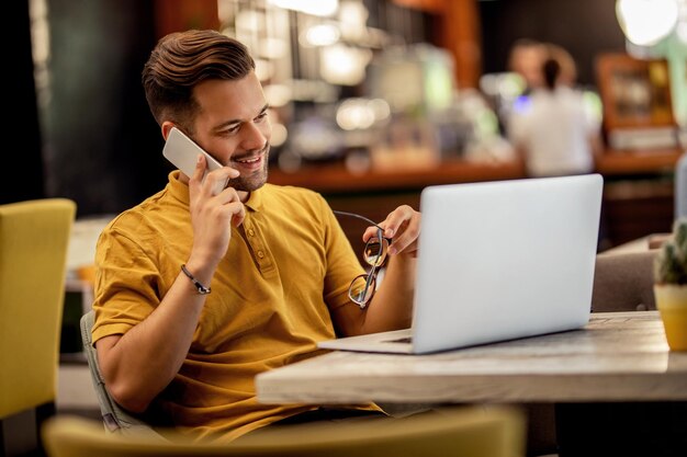 Joven trabajador independiente feliz trabajando en una computadora mientras habla por teléfono inteligente en un bar