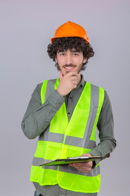 Joven trabajador ingeniero guapo empanizado de aspecto inteligente tocando su barbilla sonriendo de pie sobre fondo blanco aislado