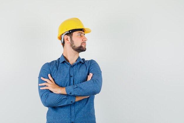 Joven trabajador mirando a un lado con los brazos cruzados en camisa, casco y mirando enfocado.