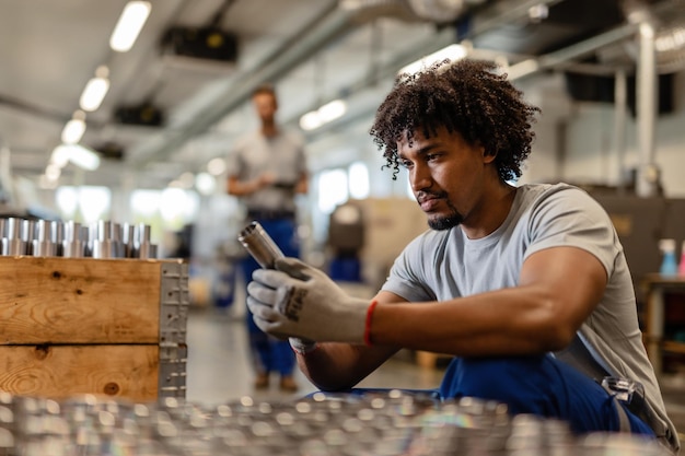 Joven trabajador negro que examina la varilla del cilindro de acero inoxidable mientras trabaja en el almacén de distribución