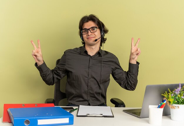 Joven trabajador de oficina complacido en auriculares con gafas ópticas se sienta en el escritorio con herramientas de oficina usando laptop y gestos signo de mano de victoria aislado sobre fondo verde con espacio de copia