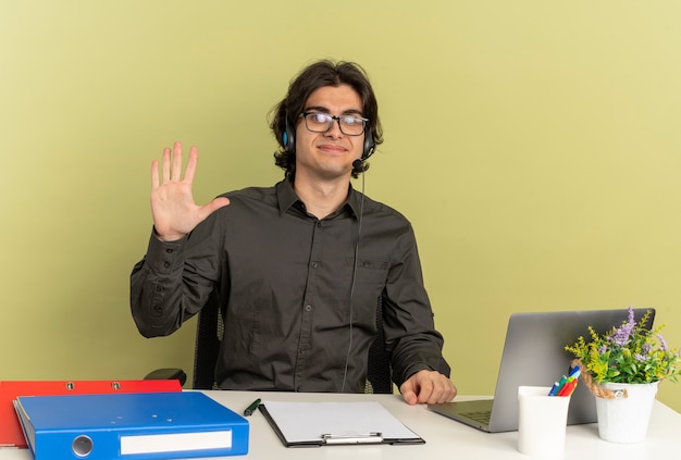 Joven trabajador de oficina complacido en auriculares con gafas ópticas se sienta en el escritorio con herramientas de oficina usando laptop levanta la mano mirando a cámara aislada sobre fondo verde con espacio de copia
