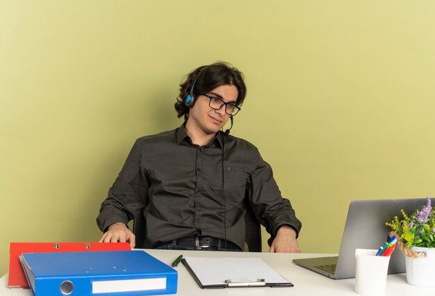 Joven trabajador de oficina complacido en auriculares con gafas ópticas se sienta en el escritorio con herramientas de oficina usando y mirando portátil aislado sobre fondo verde con espacio de copia