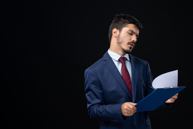 Joven trabajador de oficina masculino orgulloso en traje sosteniendo documentos y verificando información en una pared oscura aislada