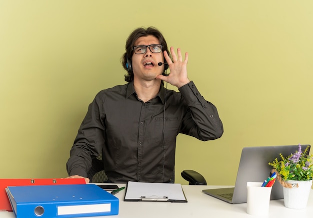 Foto gratuita joven trabajador de oficina molesto en auriculares se sienta en el escritorio con herramientas de oficina usando laptop pretendiendo llamar a alguien aislado sobre fondo verde con espacio de copia