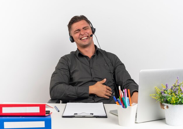 Joven trabajador de oficina rubia alegre en auriculares se sienta en el escritorio con herramientas de oficina usando laptop sostiene el vientre con los ojos cerrados