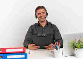 Foto gratuita joven trabajador de oficina rubia alegre en auriculares se sienta en el escritorio con herramientas de oficina usando laptop tiene portapapeles aislado sobre fondo blanco con espacio de copia