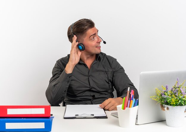 Joven trabajador de oficina rubia complacido en auriculares se sienta en el escritorio con herramientas de oficina usando laptop mira al lado con la mano levantada aislada sobre fondo blanco con espacio de copia