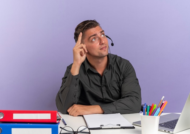 Foto gratuita joven trabajador de oficina rubia complacido en auriculares se sienta en el escritorio con herramientas de oficina usando puntos de portátil y mira hacia arriba