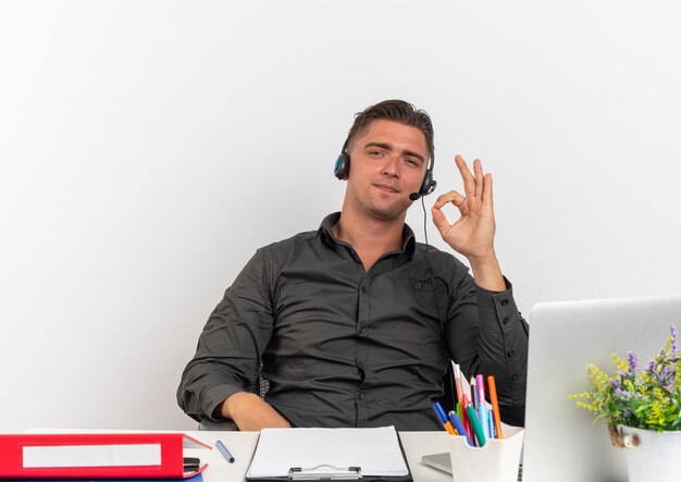 Joven trabajador de oficina rubia confía en auriculares se sienta en el escritorio con herramientas de oficina usando gestos de laptop signo de mano ok mirando a cámara aislada sobre fondo blanco con espacio de copia