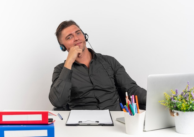 Foto gratuita joven trabajador de oficina rubia confía en auriculares se sienta en el escritorio con herramientas de oficina usando laptop pone la mano en la barbilla aislada sobre fondo blanco con espacio de copia