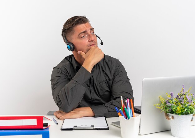 Joven trabajador de oficina rubia pensativa en auriculares se sienta en el escritorio con herramientas de oficina usando laptop pone la mano en la barbilla mirando a un lado