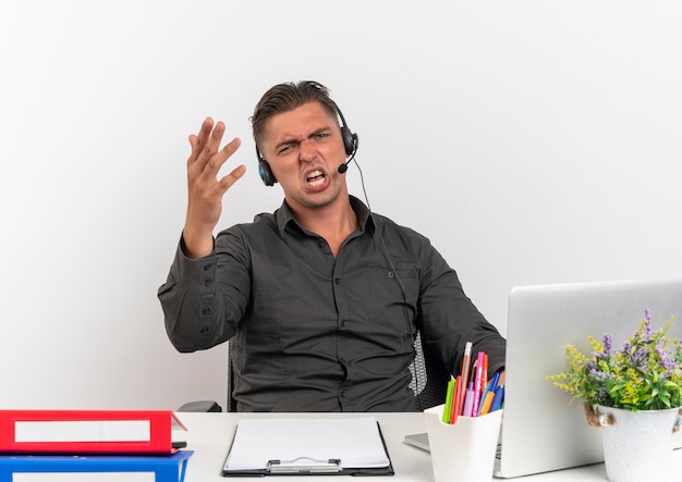 Joven trabajador de oficina rubio molesto en auriculares se sienta en el escritorio con herramientas de oficina usando gritos de computadora portátil con la mano levantada aislada sobre fondo blanco con espacio de copia