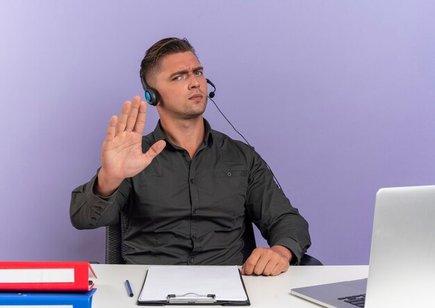 Joven trabajador de oficina rubio serio en auriculares se sienta en el escritorio con herramientas de oficina usando gestos de portátil dejar de firmar la mano aislada sobre fondo violeta con espacio de copia