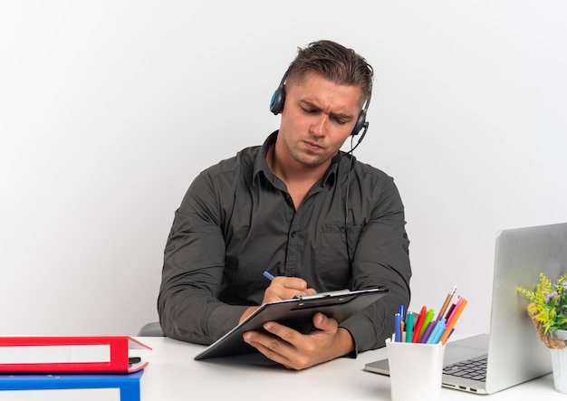 Joven trabajador de oficina rubio serio en auriculares se sienta en el escritorio con herramientas de oficina usando laptop escribe con lápiz en el portapapeles