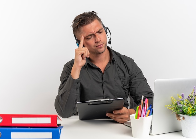 Foto gratuita joven trabajador de oficina rubio serio en auriculares se sienta en el escritorio con herramientas de oficina usando laptop sostiene y mira el portapapeles