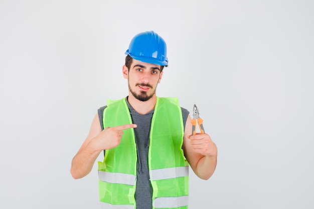 Joven trabajador sosteniendo alicates y apuntando hacia él en uniforme de construcción y mirando feliz