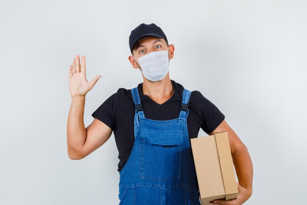 Joven trabajador sosteniendo una caja de cartón y agitando la mano en uniforme, vista frontal de la máscara.