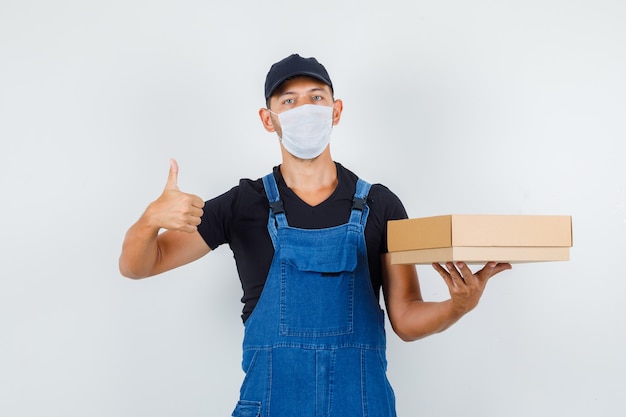 Joven trabajador sosteniendo una caja de cartón con el pulgar hacia arriba en uniforme, vista frontal de la máscara.