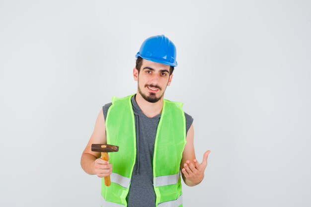 Joven trabajador sosteniendo el hacha en una mano mientras estira la otra mano mientras sostiene algo en uniforme de construcción y se ve feliz