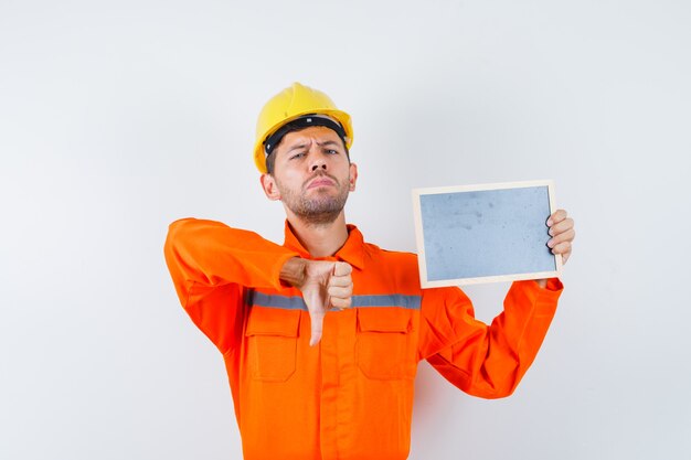 Joven trabajador sosteniendo la pizarra, mostrando el pulgar hacia abajo en uniforme, casco y mirando descontento.