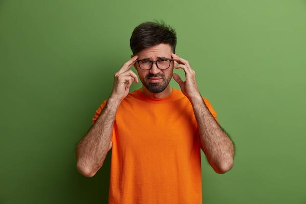 El joven trabajador tiene un dolor de cabeza insoportable, mantiene las manos en las sienes, frunce el ceño por el dolor, siente migraña dolorosa, trabaja con exceso de trabajo durante la preparación del proyecto, usa gafas transparentes y una camiseta naranja