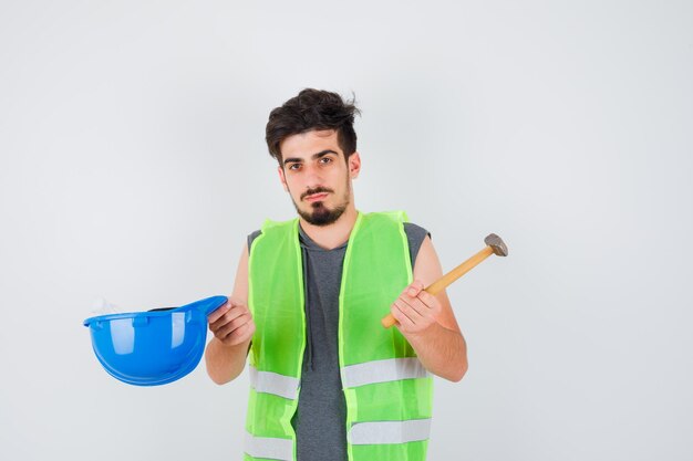 Joven trabajador en uniforme de construcción sosteniendo el hacha en una mano y la gorra en la otra mano y mirando serio