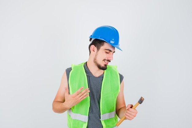 Joven trabajador en uniforme de construcción sosteniendo el hacha en una mano y mirándola y mirando feliz