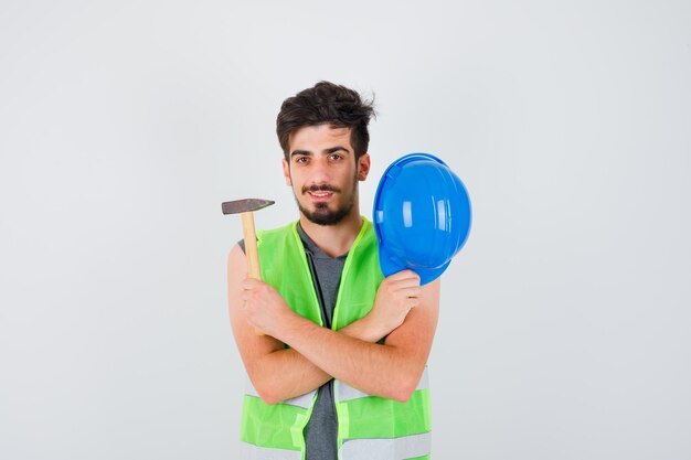 Joven trabajador en uniforme de construcción sosteniendo el hacha en una mano y quitándose la gorra y mirando feliz