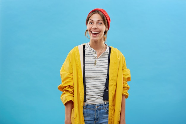Joven trabajadora bonita con chaqueta amarilla, overoles de jean y sombrero rojo posando contra la pared azul con una sonrisa feliz de terminar su trabajo sobre la casa. Emociones, concepto de felicidad
