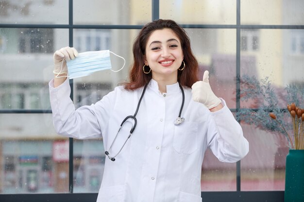 Una joven trabajadora de la salud que sostiene una máscara y un gesto con el pulgar uo Foto de alta calidad