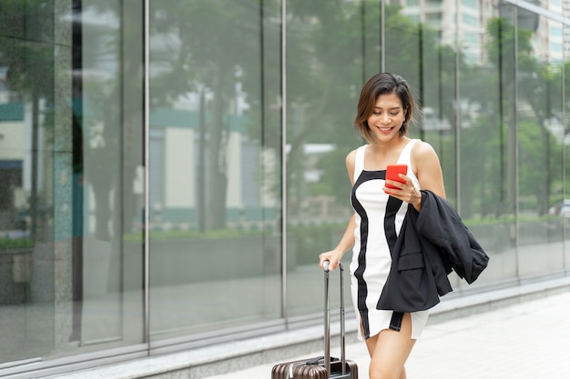 Joven trabajadora usar teléfono inteligente y caminar con maleta